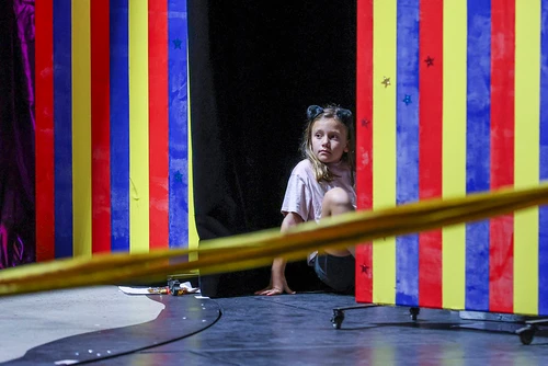 Ein Kind hinter einer bunten Rollwand sitzend auf die Bühne schauend.