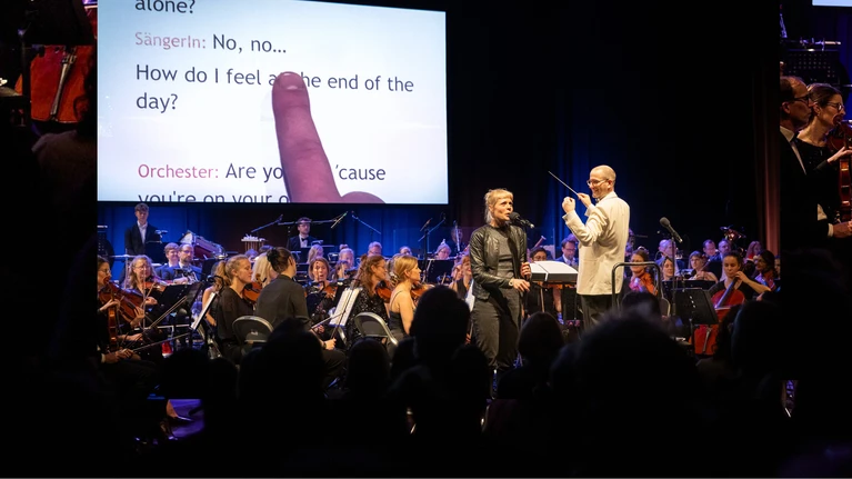 Foto von "Orchesterkaraoke", auf der Bühne ein Orchester, dahinter wird offenbar ein Songtext projiziert, eine Person steht an einem Mikrofon und singt.