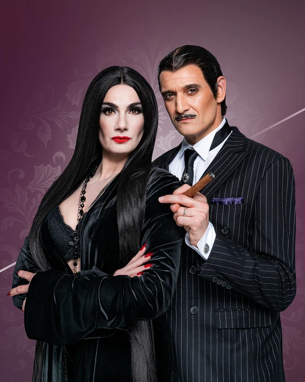 Two people pose as Morticia and Gomez Addams in classic costumes against a dark purple floral background.