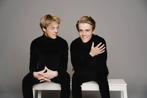 zwei blonde junge Männer, Geschwister, in dunklen Anzügen sitzen auf einer weißen Bank