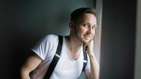 Mann in weißem T-Shirt und Hosenträgern sitzt an einem Fenster und stützt sich auf seine linke Hand