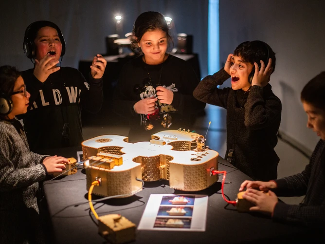 Photo of children standing around a novel, technically sophisticated-looking instrument or trying it out – ‘Mobile Touch’, a sound project from the BIG BANG Festival 2026.