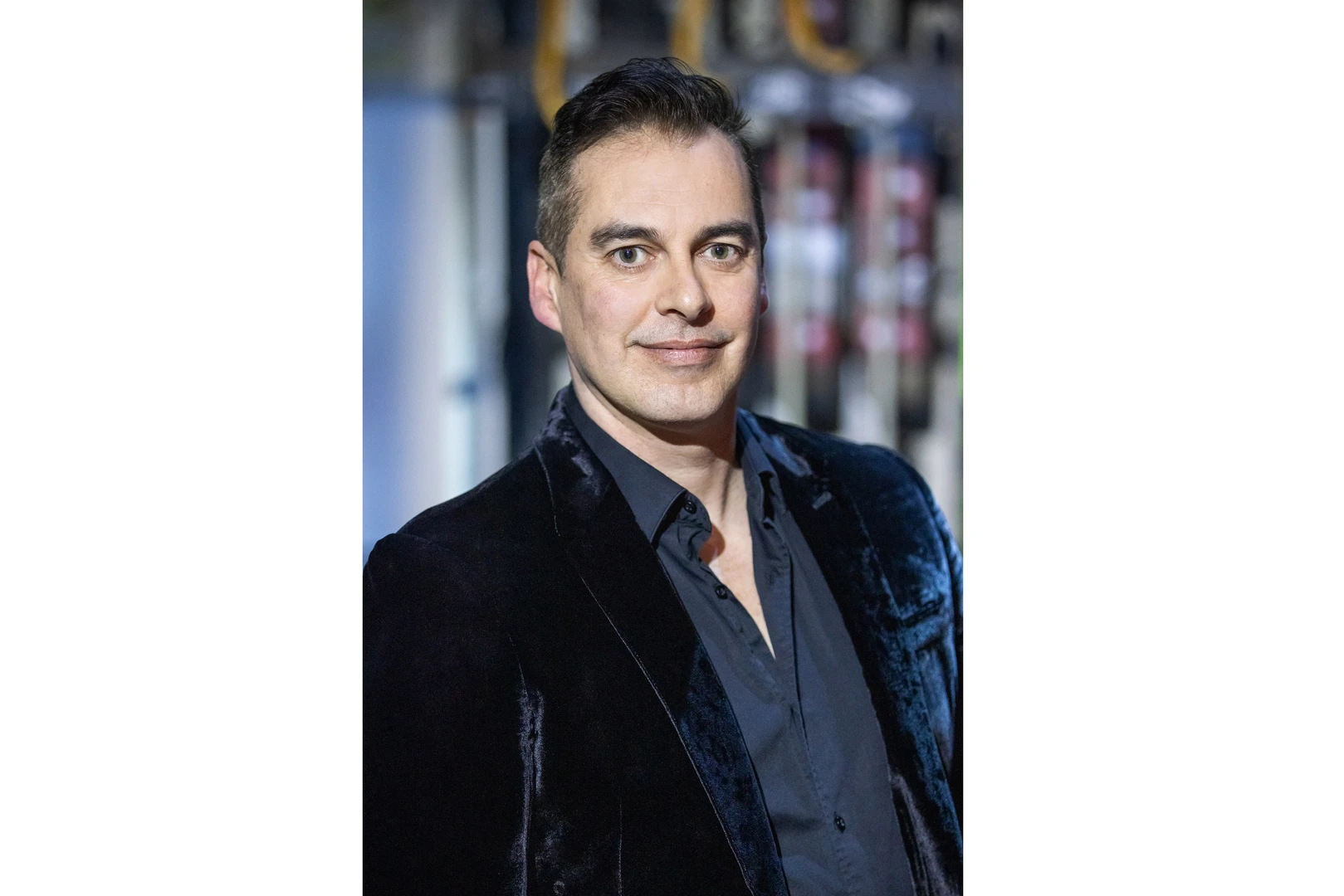 Eine Person trägt ein dunkles Samt-Sakko über einem dunklen Hemd; der Hintergrund zeigt verschwommene Studioelemente. Das Gesicht ist absichtlich unscharf und nicht erkennbar.