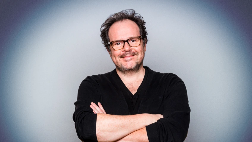 Thomas Mraz steht lächelnd mit Brille und dunkler Kleidung vor einem neutralen grauen Hintergrund und hat die Arme locker vor der Brust verschränkt.