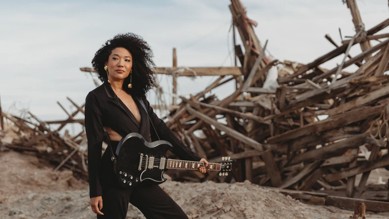 Pressefoto von Judith Hill im schwarzen Hosenanzug mit E-Gitarre vor einem Schiffswrack aus Holz