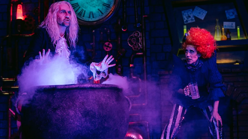 Eine Person mit langen weißen Haaren hält die Hände über einem schwarzen, dampfenden Kessel, daneben eine Person mit roten lockigen Haaren und einem Glas.