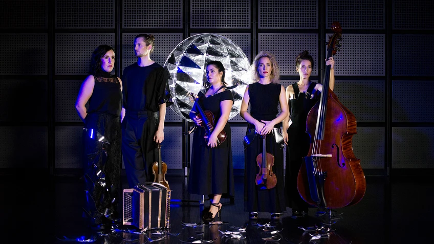 Die fünf Musiker:innen von ALMA stehen in eleganten schwarzen Outfits vor einer metallisch glänzenden, kuppelförmigen Lichtinstallation, halten ihre Instrumente – Harmonium, Violinen, Bratsche und Kontrabass – und blicken ernst in die Kamera.