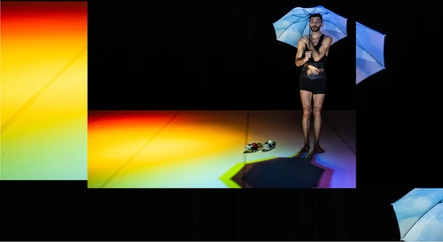Photo from the performance ‘Souvenirshop’ performed by BRONKS & Not Standing, choreographed by Alexander Vantournhout. It shows a man dressed in black holding an umbrella on an almost completely dark stage – light refracts on the floor, forming a rainbow.