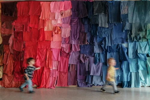 Materiallabor "Glatt oder rau?". Kinder spielen vor einer Wand aus bunten Stoffen, die wie ein Regenbogen sortiert sind.