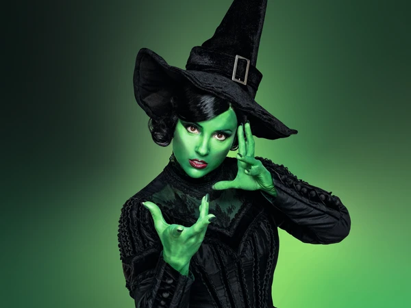 Green background. Woman with green colored face and pointed black hat as well as black dress looks into the camera, her hands spread in front of her face. Close-up.