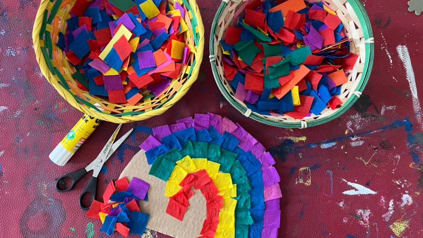 Zwei Körbe mit bunten Papierstücken, daneben eine Schere, ein Klebestift und eine Regenbogen‑Collage auf bemaltem Tisch.