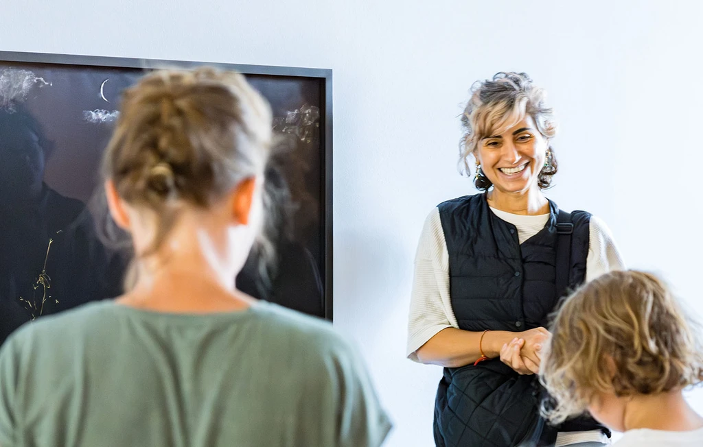 Künstlerin Nilbar Güreş steht in der Ausstellung "Papier, Stein, Schere" mit Workshopteilnehmer:innen vor ihrer textilen Collage "Picknick im Dunklen".