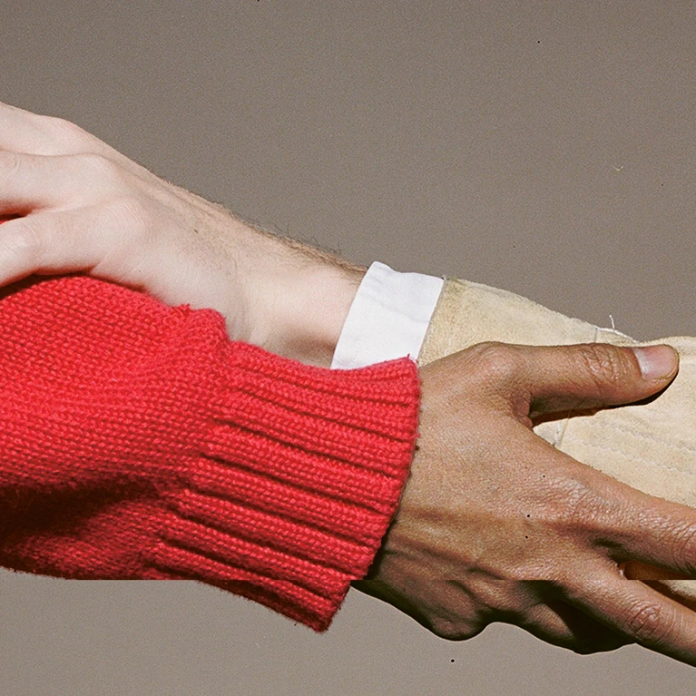 The theme for the Festspielhaus’s 2026/2027 season. The image shows hands touching, or rather two hands touching each other’s arms in a supportive gesture; one person’s sleeve is a deep red, the other’s a light beige, set against a dark beige background.
