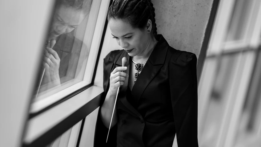 Frau mit Dirigierstock in der Hand, ihr Blickt geht nach unten, neben einer Glasfensterfront