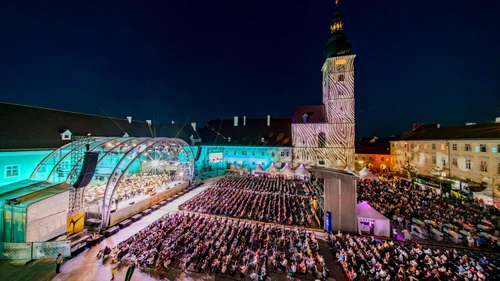 Großer Platz mit einer Bühne und viel Publikum davor in einer Stadt am Abend