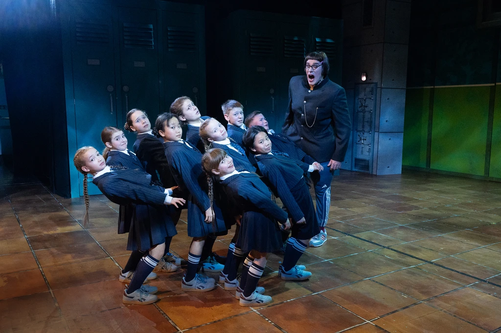 A group of children in dark school uniforms recoil in fear from a large person who is shouting at them. Tiled floor, lockers in the background, cool and dramatic lighting.