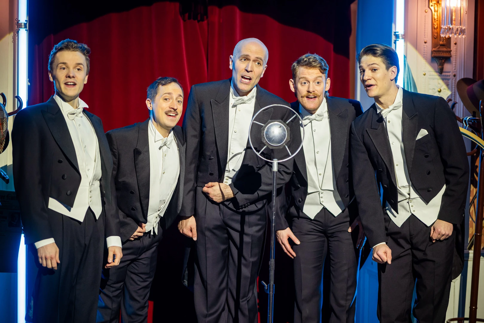 Five men in tailcoats, singing behind an old-fashioned microphone, with a red velvet curtain in the background