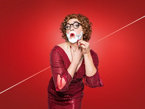 Eine Person in rotem Glitzerkleid mit Perlenkette und Locken rasiert sich das Gesicht vor rotem Hintergrund.
