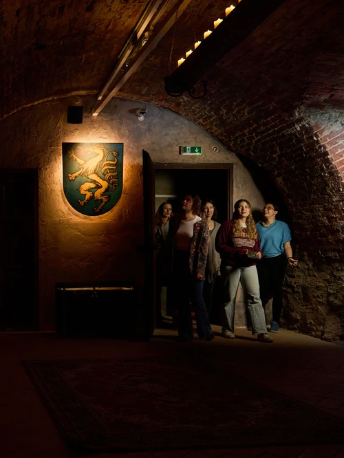 5 Frauen betreten ein Kellergewölbe mit einem Goldenen-Panther-Wappen.