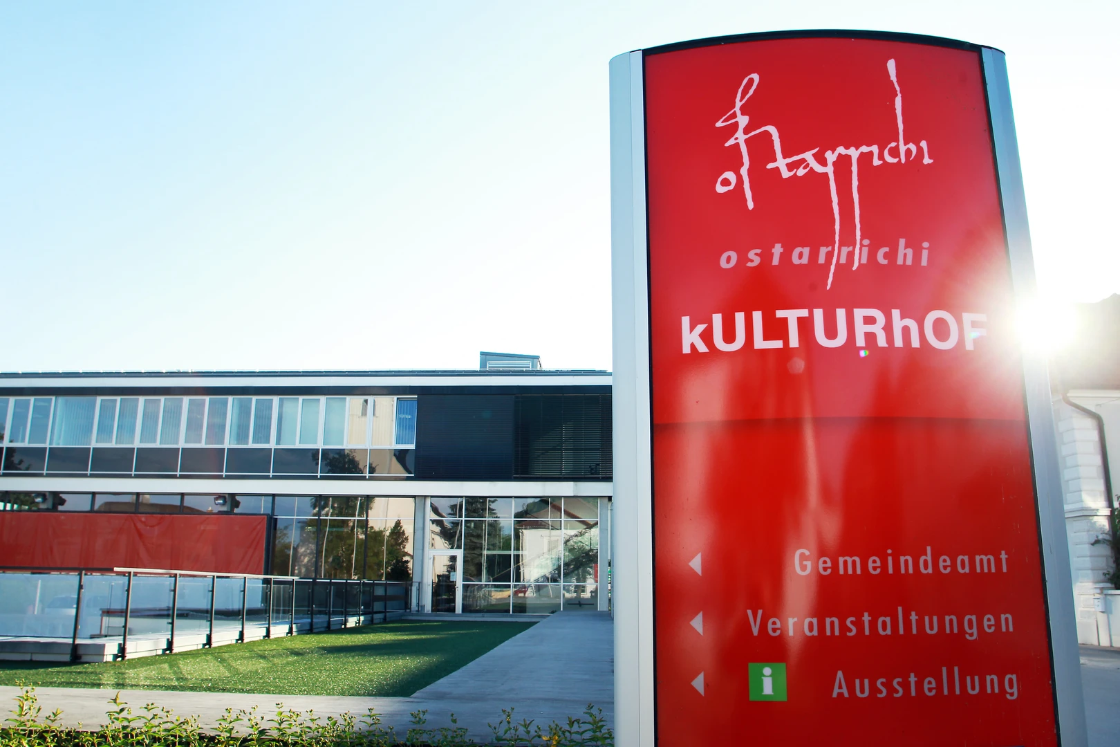 Außenansicht des Museums Ostarrichi mit roter Tafel ‚Ostarrichi‘ und Hinweisen zu Kulturhof, Gemeindeamt, Veranstaltungen und Ausstellung, daneben das moderne Gebäude mit großer Glasfront, schwarzer Glasfassade im oberen Stockwerk, asphaltierter Fläche und grüner Wiese.