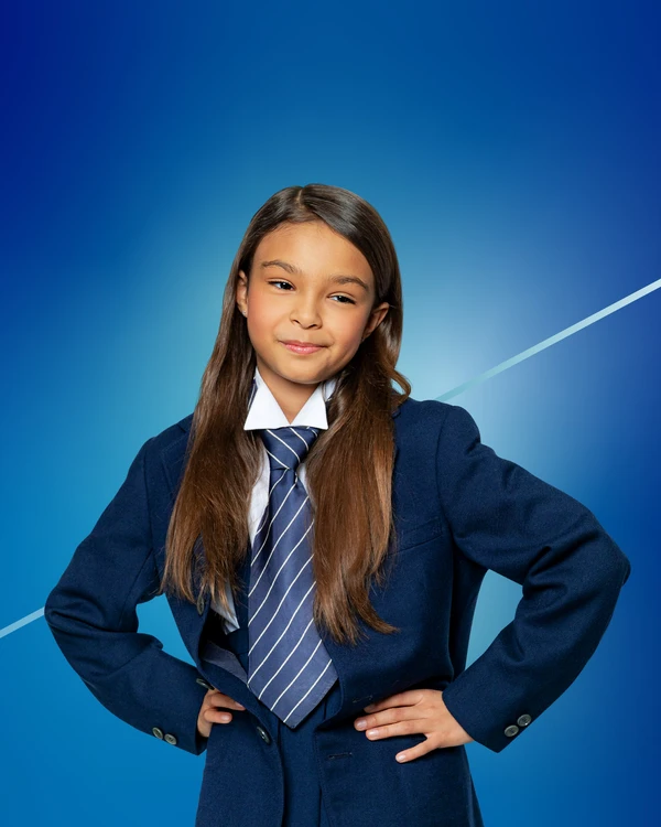Ein circa neunjähriges, braunhaariges Mädchen in einer dunkelblauen Schuluniform mit Krawatte steht mit Händen in die Hüften gestützt vor einem blauen Hintergrund. Nahaufnahme.