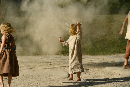 Two children in tunics are playing in the street; the child in the middle is throwing sand into the air 