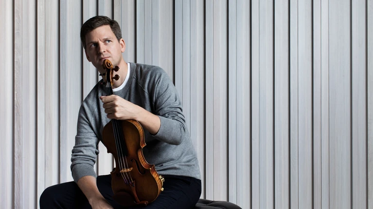 Photograph of violinist James Ehrens, seen with his instrument, seated on a stool in front of a light grey curtain.