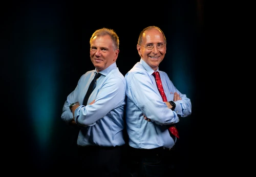Foto von Journalist und ORF-Moderator Armin Wolf und der ebenfalls durch den ORF bekannte Politikwissenschaftler Peter Filzmaier stehen – beide mit hellblauem Hemd und Krawatte – Rücken an Rücken aneinander vor einem schwarzen Bildhintergrund und lächeln seitlich zur Kamera.