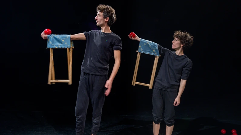 Zwei Künstler:innen in schwarzer Kleidung balancieren jeweils einen kleinen Hocker auf ihrem ausgestreckten Arm, während sie rote Jonglierbälle halten und die Wand auf einer dunklen Bühne konzentriert anschauen.