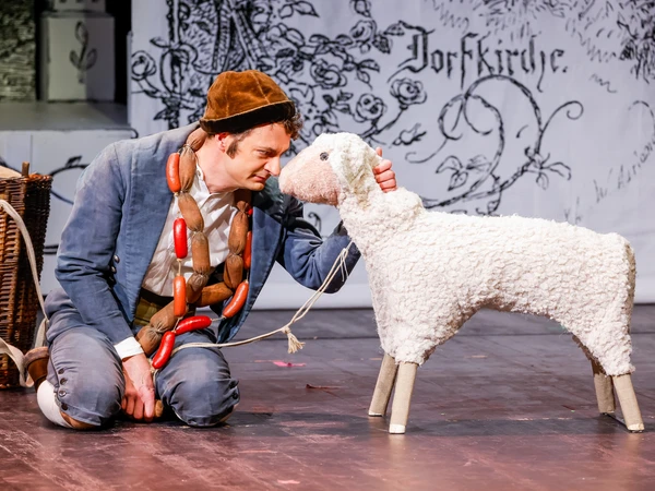 Man with a sausage chain around his neck kneeling in front of a sheep, to his left is a basket, in the background a white wall with a black floral pattern