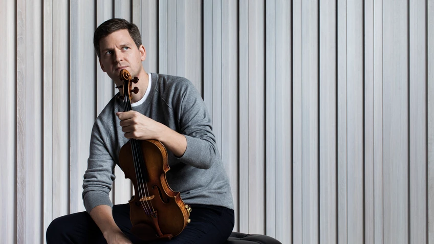 Photograph of violinist James Ehrens, seen with his instrument, seated on a stool in front of a light grey curtain.