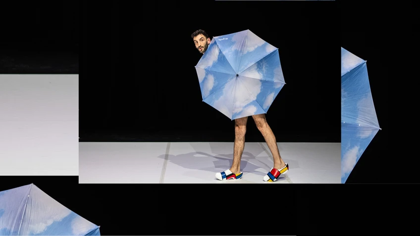 Foto vom Stück "Souvenirshop" - zu sehen ist ein Performer, der einen Regenschirm vor sich hält, der mit einer Ansicht vom Himmel bedruckt ist. Er trägt Croc-Schuhe im Design des Künstlers Mondrian und seinen Farbflächen.
