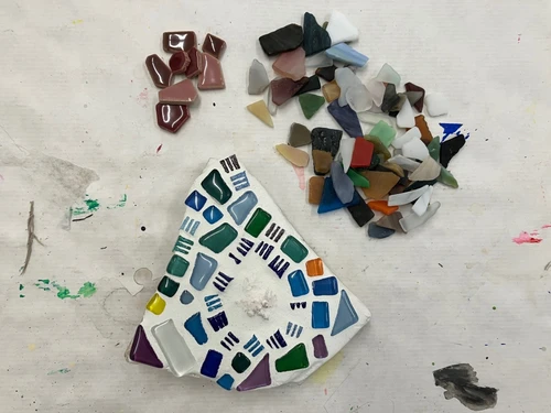 Dreieckiges Mosaik aus bunten Glasstücken auf einem Tisch, daneben lose Scherben in verschiedenen Farben und Formen.