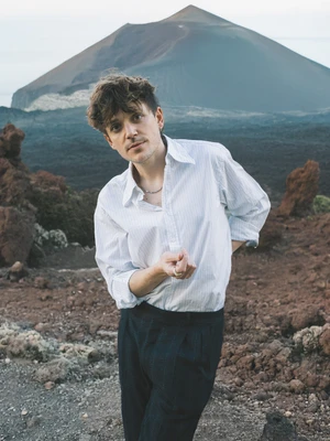 Foto von Ariel Oehl, dem Musiker hinter dem Musikprojekt "Oehl" in weißem Hemd und schwarzer Hose vor einer kargen Landschaft, im Hintergrund ein hoher Berg.