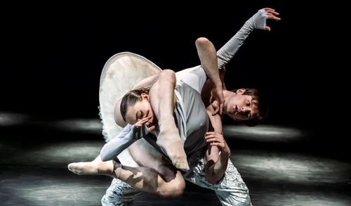 Zwei Tänzer:innen performen "Mémoires d’Oubliettes", eine Choreografie von Jirí Kylián. Beide tragen weiße Kostüme und sind so miteinander körperlich verwoben, dass ihr Gliedmaßen kaum mehr einer Person zuordbar sind.
