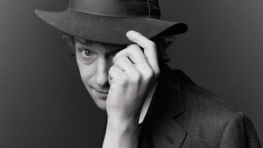 Porträtfoto des Musikers Voodoo Jürgens in schwarz-weiß, der mit auffälligem Hut Richtung Kamera grüßt.