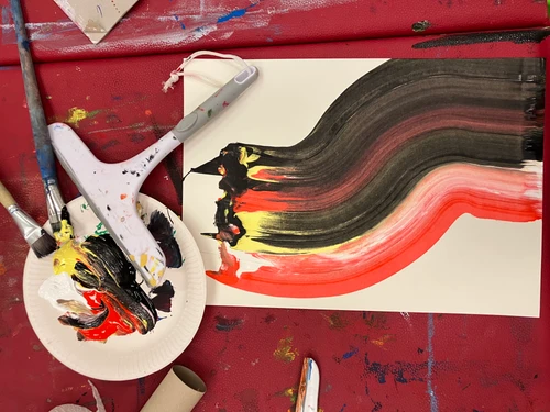 Pinsel, Scheibenwischer und Farben liegen auf einem Tisch. Das Blatt Papier wurde angemalt mit mehreren Farben die ineinander vermischt sind.