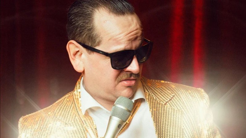 Romantic Slivo steht mit dunkler Sonnenbrille und Schnurrbart vor einem roten Bühnenvorhang, trägt einen auffälligen goldenen Glitzeranzug und hält ein Mikrofon mit Kabel in der Hand, während er seitlich nach unten blickt.