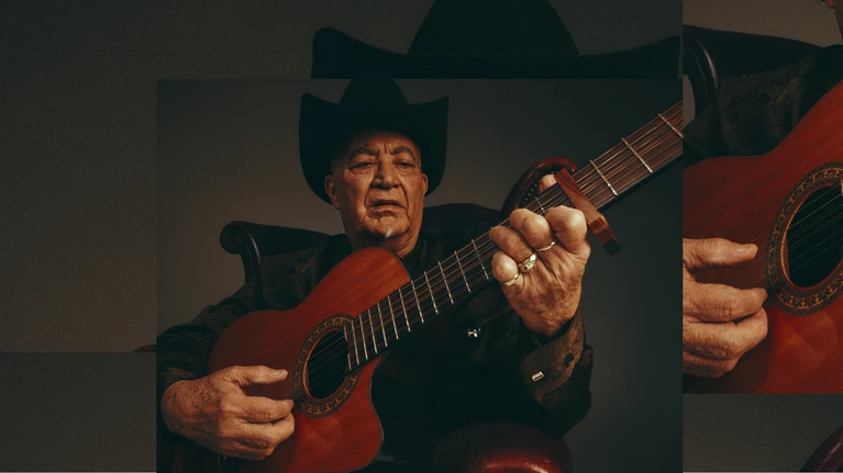 A portrait photograph of musician Elias Ochoa with his instrument, an acoustic guitar, wearing a black suit and a black hat.