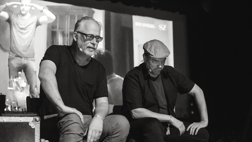 Peter Hörmanseder und Robert Stachel sitzen auf einer dunklen Bühne mit amüsiertem und ernstem Ausdruck nebeneinander, während hinter ihnen auf einer großen Leinwand eine Szene aus einer Nachrichtensendung projiziert wird.
