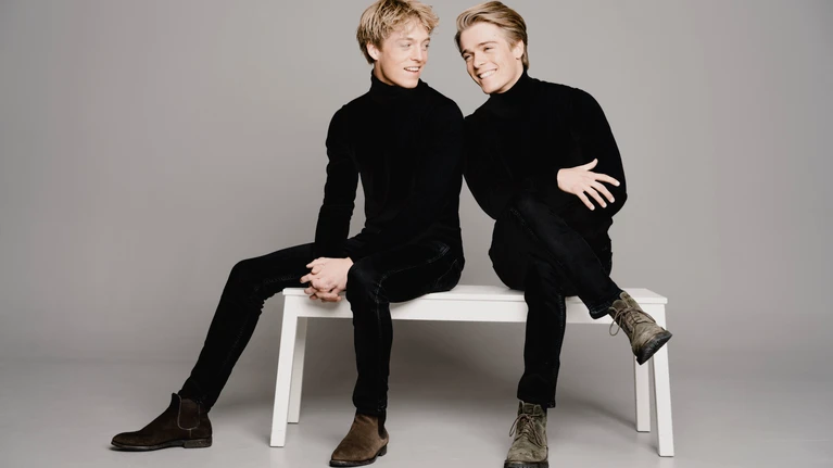 Photograph of brothers Lucas and Arthur Jussen, virtuoso pianists, seated on a white bench in front of a light grey wall.