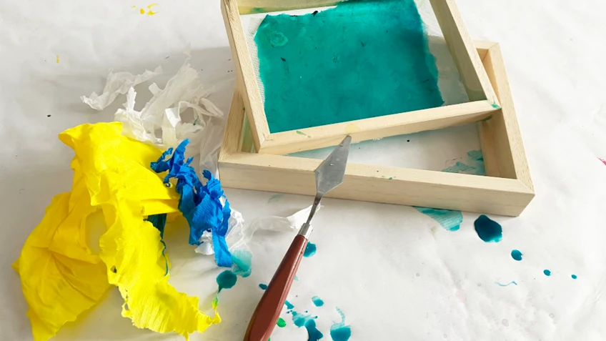 Kleiner Arbeitsbereich mit zwei leeren Holzrahmen, türkis eingefärbtem Papier, gelben und blauen Farbresten sowie einem Spachtel auf weißer Unterlage.
