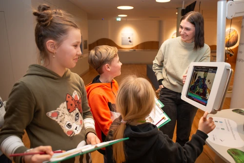 Drei Kinder haben ein Klemmbrett und nehmen an der Rätselrallye im Haus der Wildnis teil. Ein Guide erklärt ihnen die Inhalte mit einem in der Ausstellung angebrachten, interaktiven Tablet. 