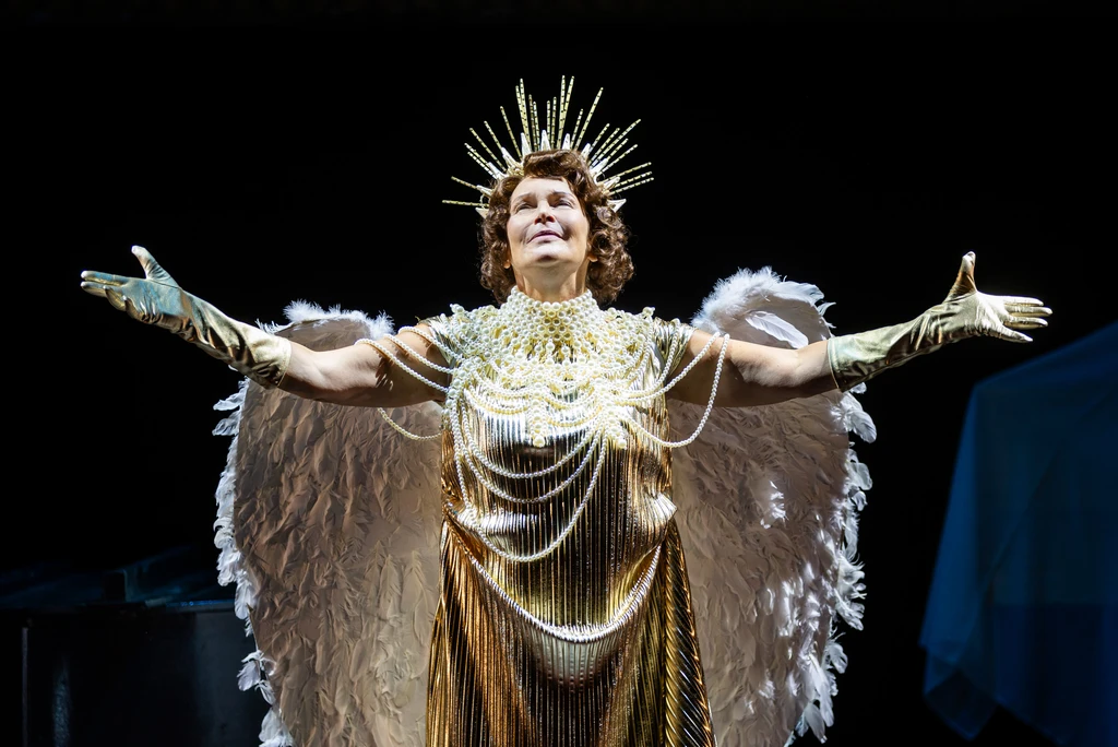 A person in a gold-coloured stage costume, with arms outstretched, stands against a dark background. The costume features large, white wings and a radiant headdress.