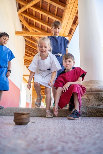 Children are playing Orca at the Porticus in the Roman Quarter; a girl is throwing a stone into a bowl; the children are wearing tunics 