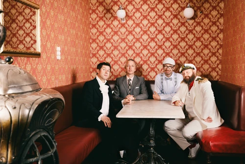 A photo of the musicians Sebastian Fuchsberger, Leonhard Paul, Michael Hornek and Thomas Gansch sitting at a table in a coffee house, with eye-catching patterned wallpaper behind them.