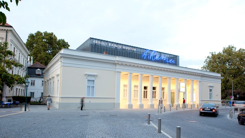 Außenansicht des Museumsgebäudes mit historischer Fassade und Säulengang vor blauem Himmel davor hellgrauer Straßenzug