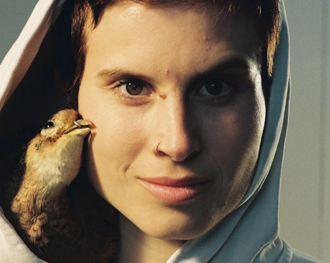 Nahaufnahme einer Frau mit dunklen kurzen Haaren, heller Hoodie mit Kapuze auf dem Kopf, ein Vogel schmiegt sich an ihr Gesicht