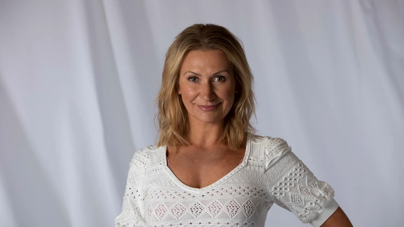 Angelika Niedetzky steht lächelnd mit schulterlangen blonden Haaren in einem weißen, gemusterten Spitzenoberteil vor einem neutralen hellen Hintergrund und blickt charmant in die Kamera.