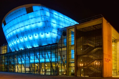 Modernes Festspielhaus mit markanter, blau beleuchteter Fassade und verglastem Eingangsbereich bei Nacht.